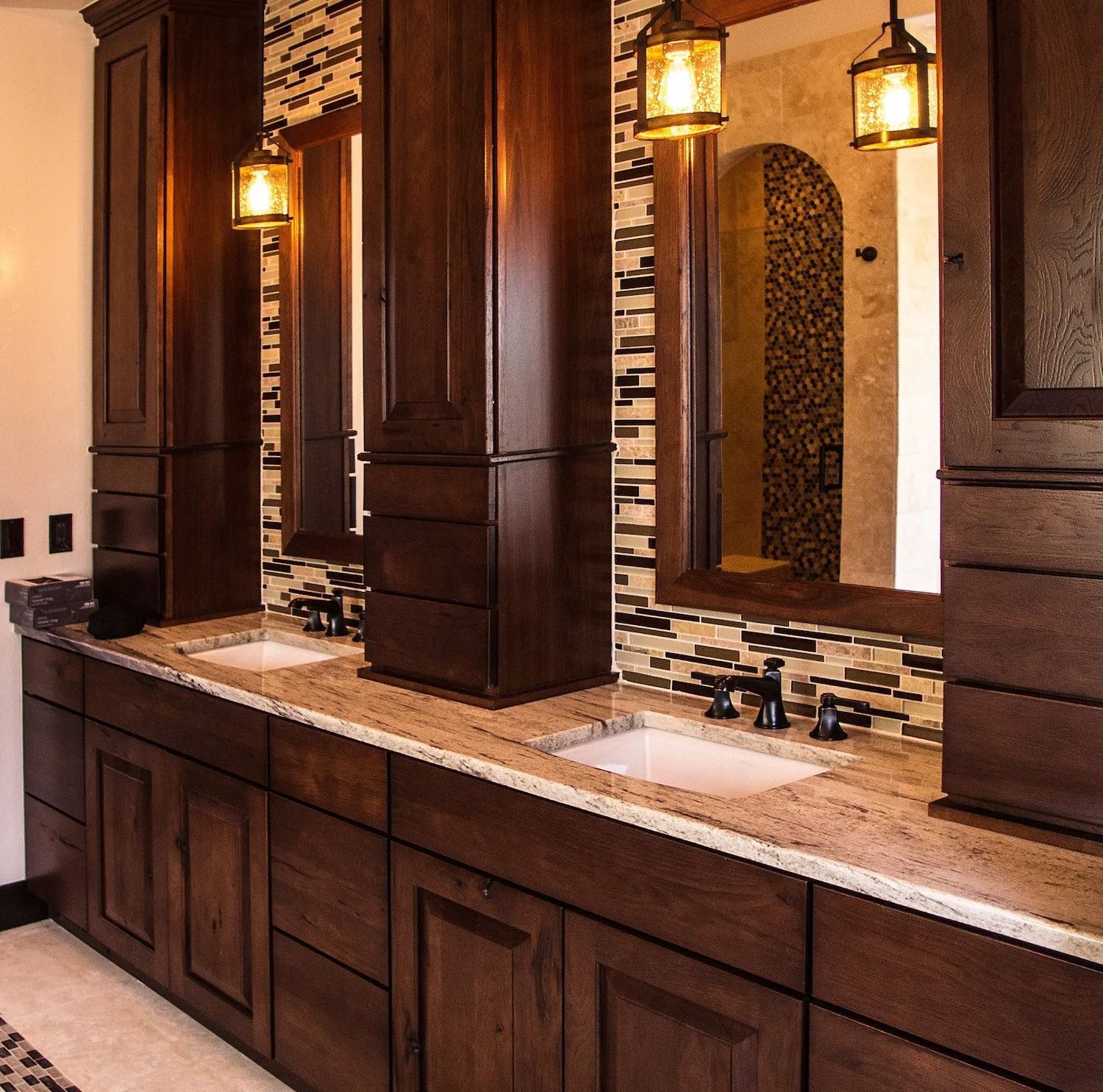 A double vanity bathroom with dark wood cabinetry, granite countertops, stone tile backsplash, and hanging pendant lights.