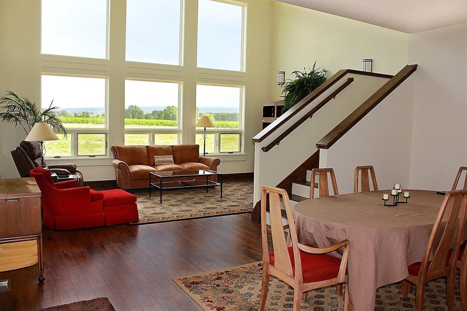A spacious living and dining area featuring a tan couch, red accent chair, large windows, and a staircase.