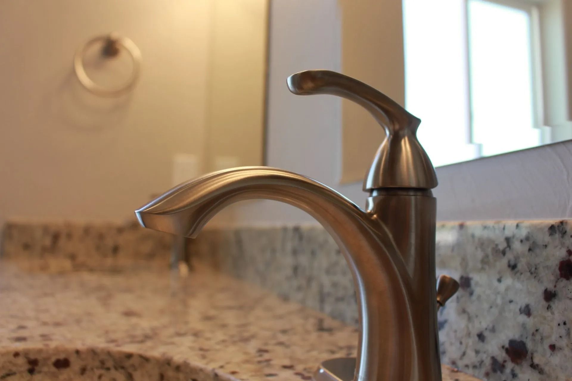 A brushed nickel bathroom faucet centered on a granite countertop, with a mirror and towel ring in the blurred background.
