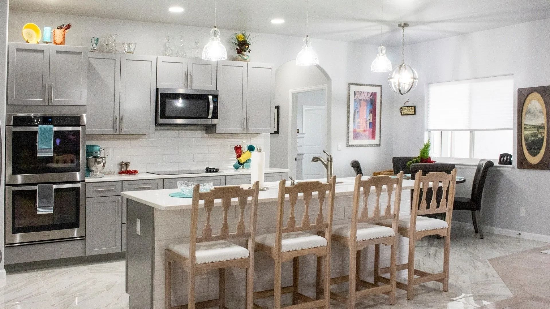 A modern kitchen with grey cabinetry, a marble-topped island with four barstools, and stainless steel appliances.