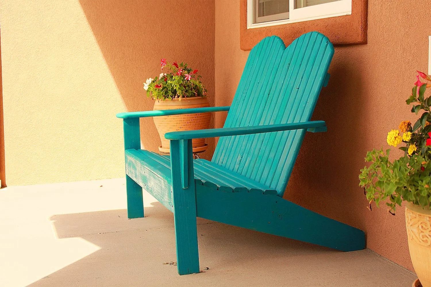 A bright turquoise wooden Adirondack-style bench sits on a patio between two potted plants against a textured wall.