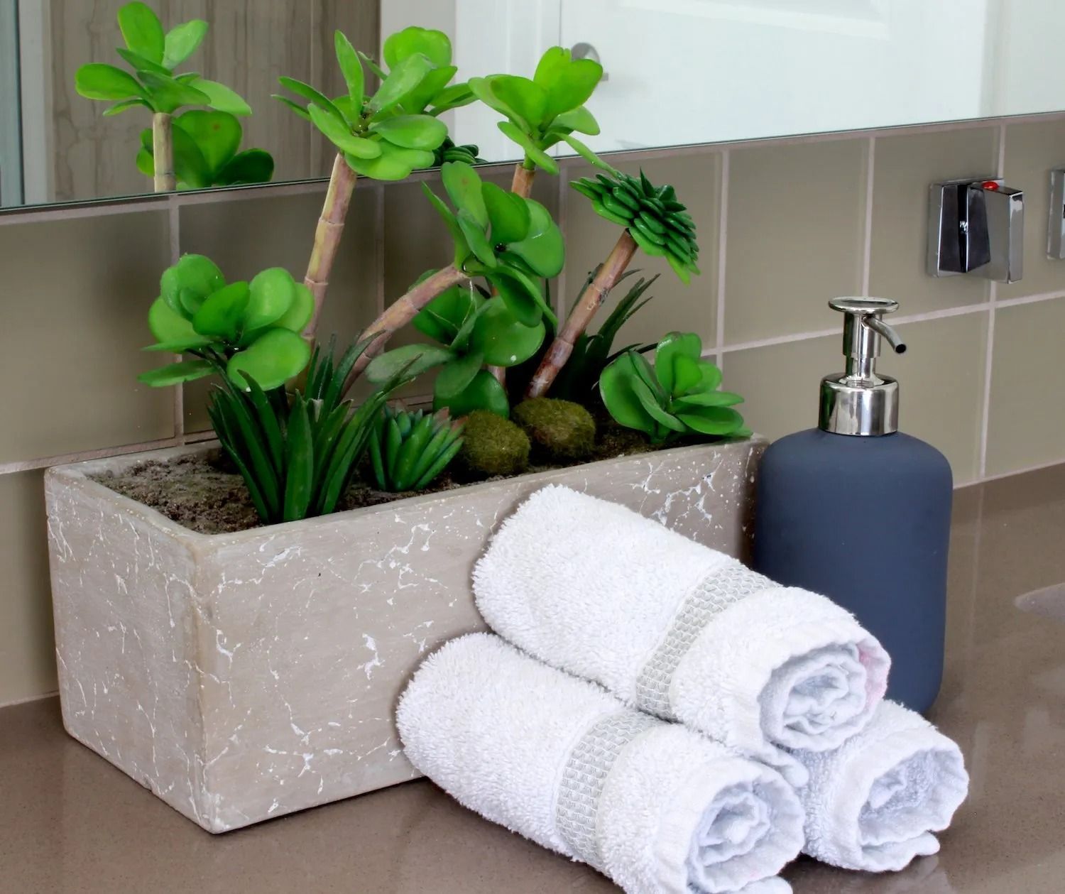 A light grey rectangular planter with artificial succulents sits next to three rolled white towels and a grey soap dispenser.