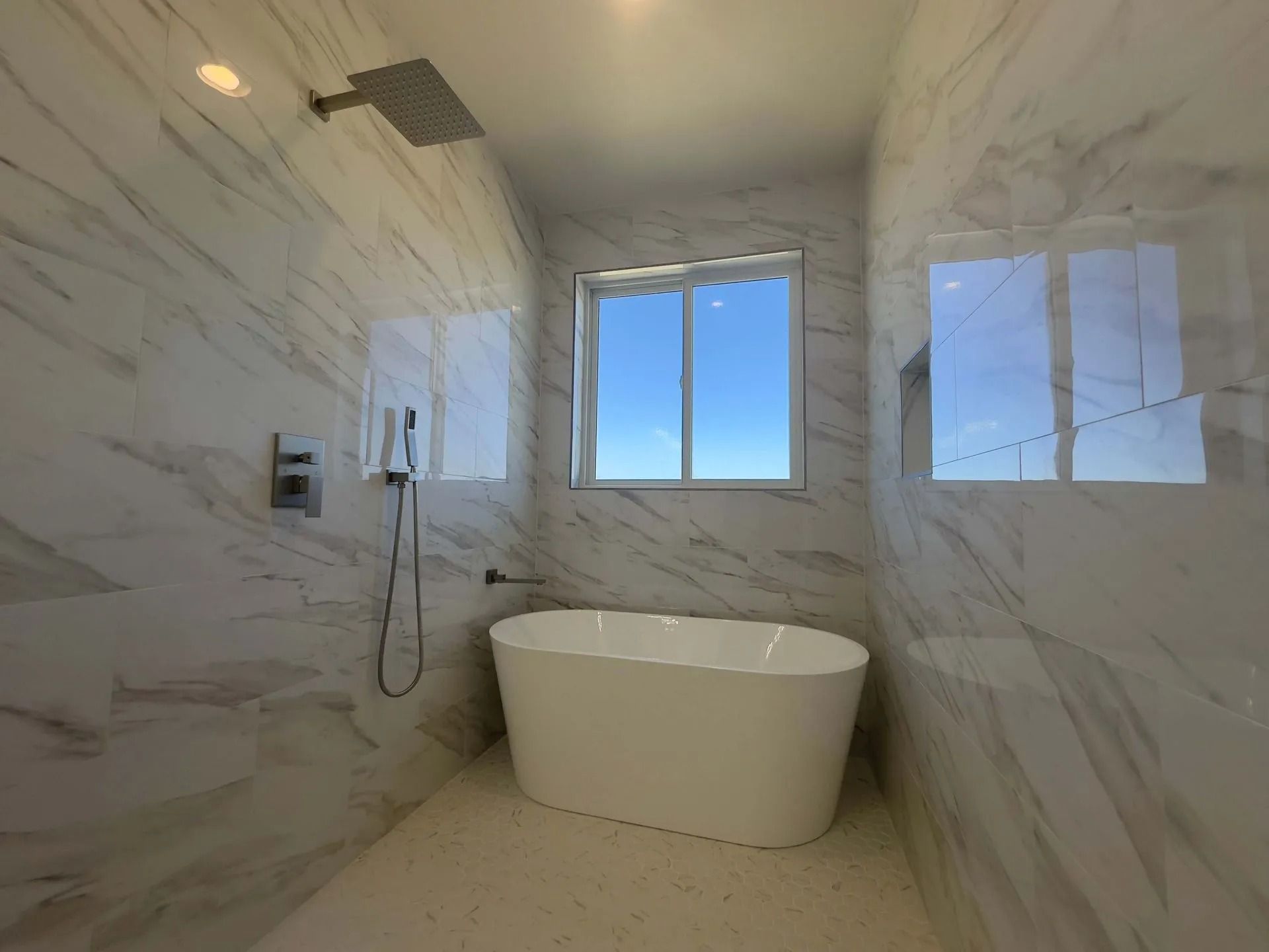A modern bathroom features a freestanding white bathtub in front of a window, surrounded by white marble-patterned walls.