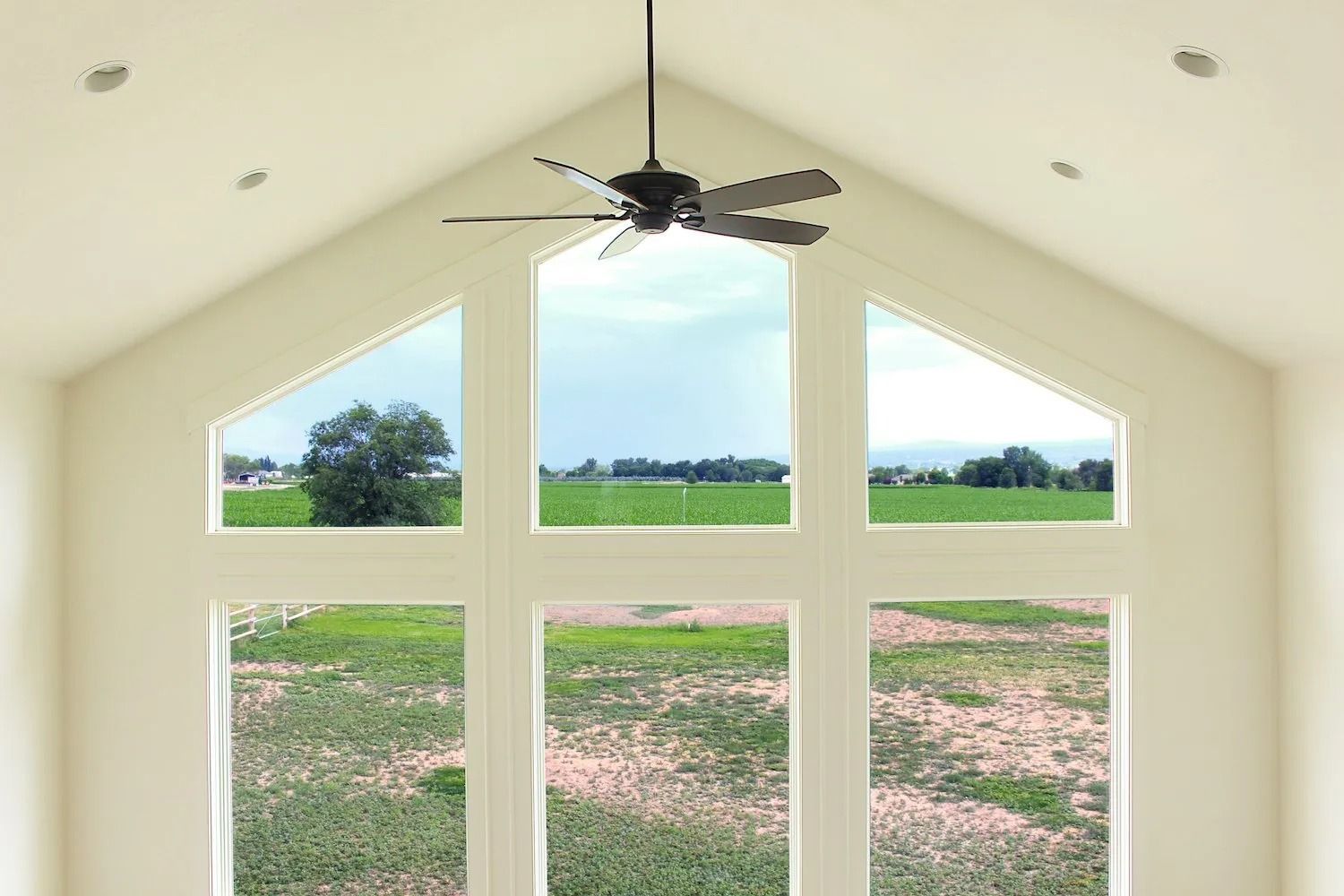 Vaulted ceiling room with a large, three-section window configuration overlooking a grassy field and a ceiling fan above.
