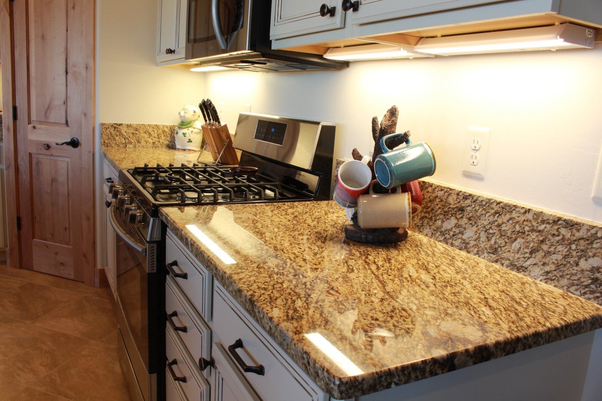 A kitchen counter with a gas stove, granite surfaces, white cabinets, and a mug tree holding several mugs.