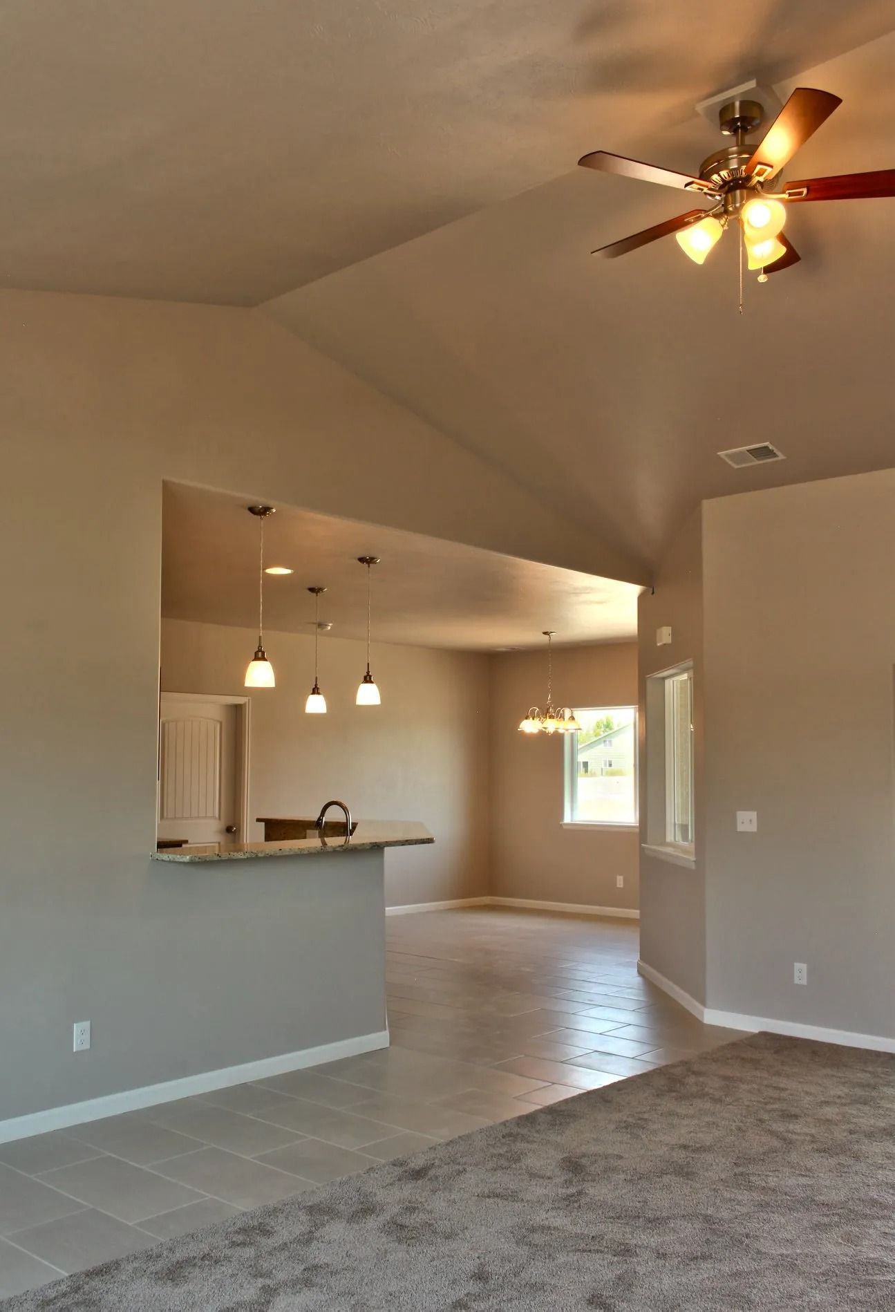 A bright, open-plan interior featuring a carpeted foreground, a kitchen with pendant lights, and a vaulted ceiling.