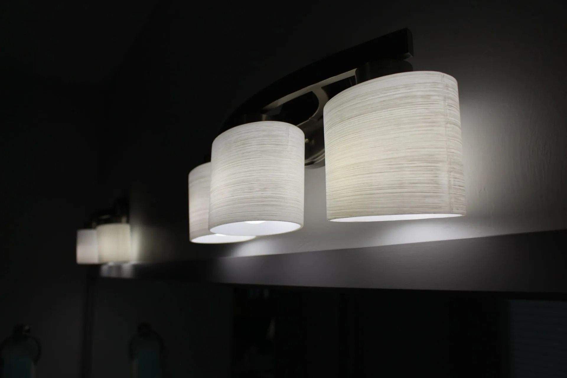 A three-light vanity fixture with textured, cylindrical white shades, reflected in a mirror against a dark wall.
