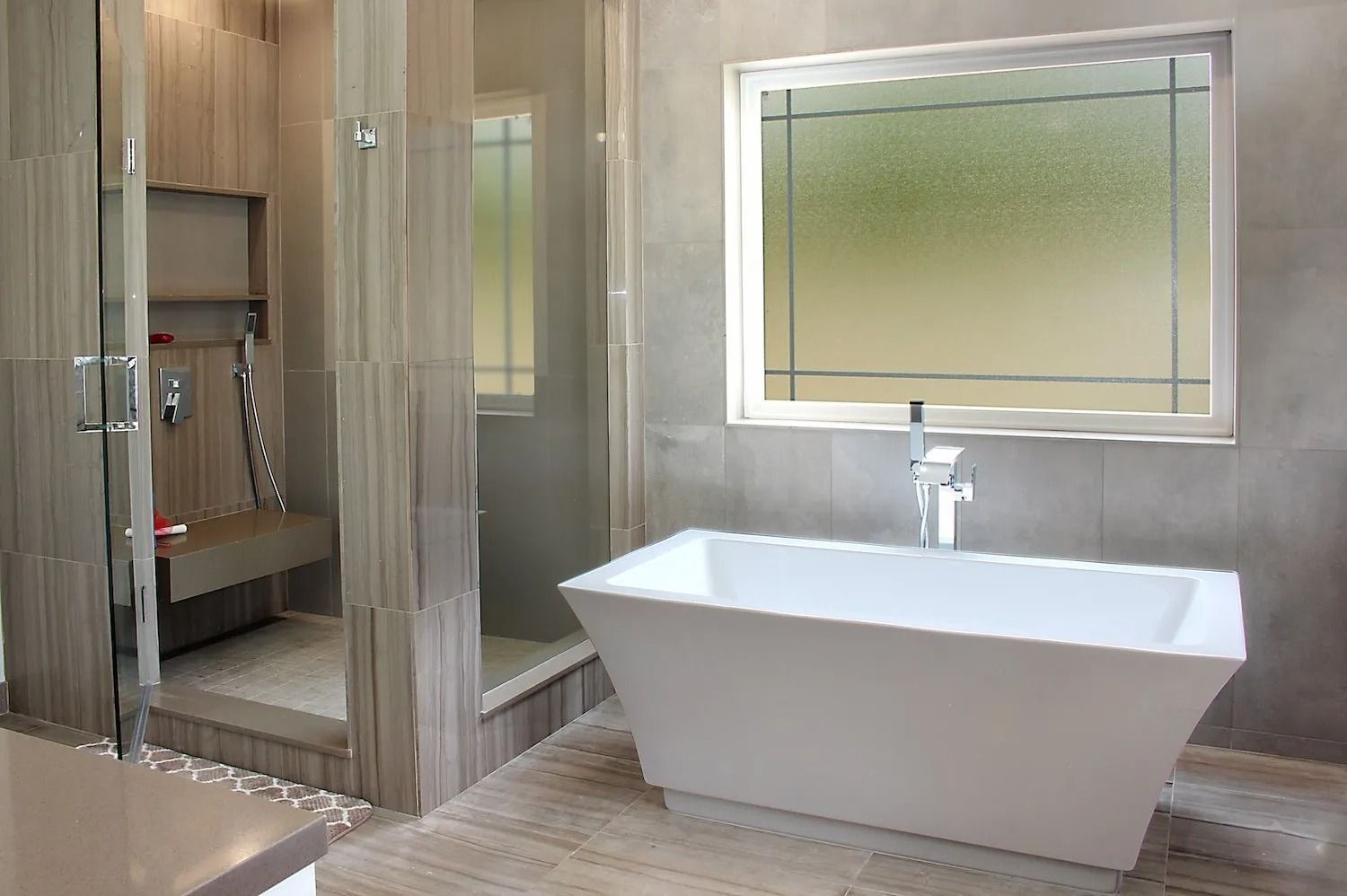 A modern bathroom with a white freestanding tub, a large window, and a walk-in glass shower with wood-tone tiling.