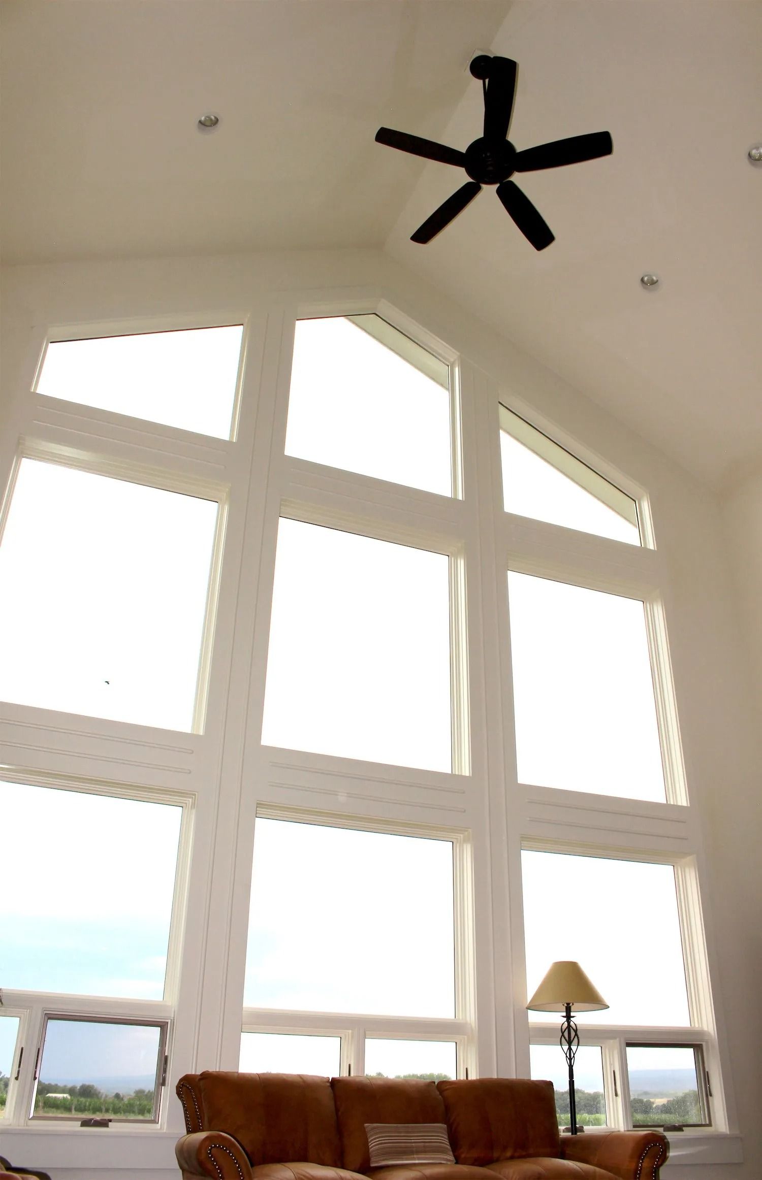 A large, white-framed window wall in a high-ceilinged room with a black ceiling fan and a brown sofa below.