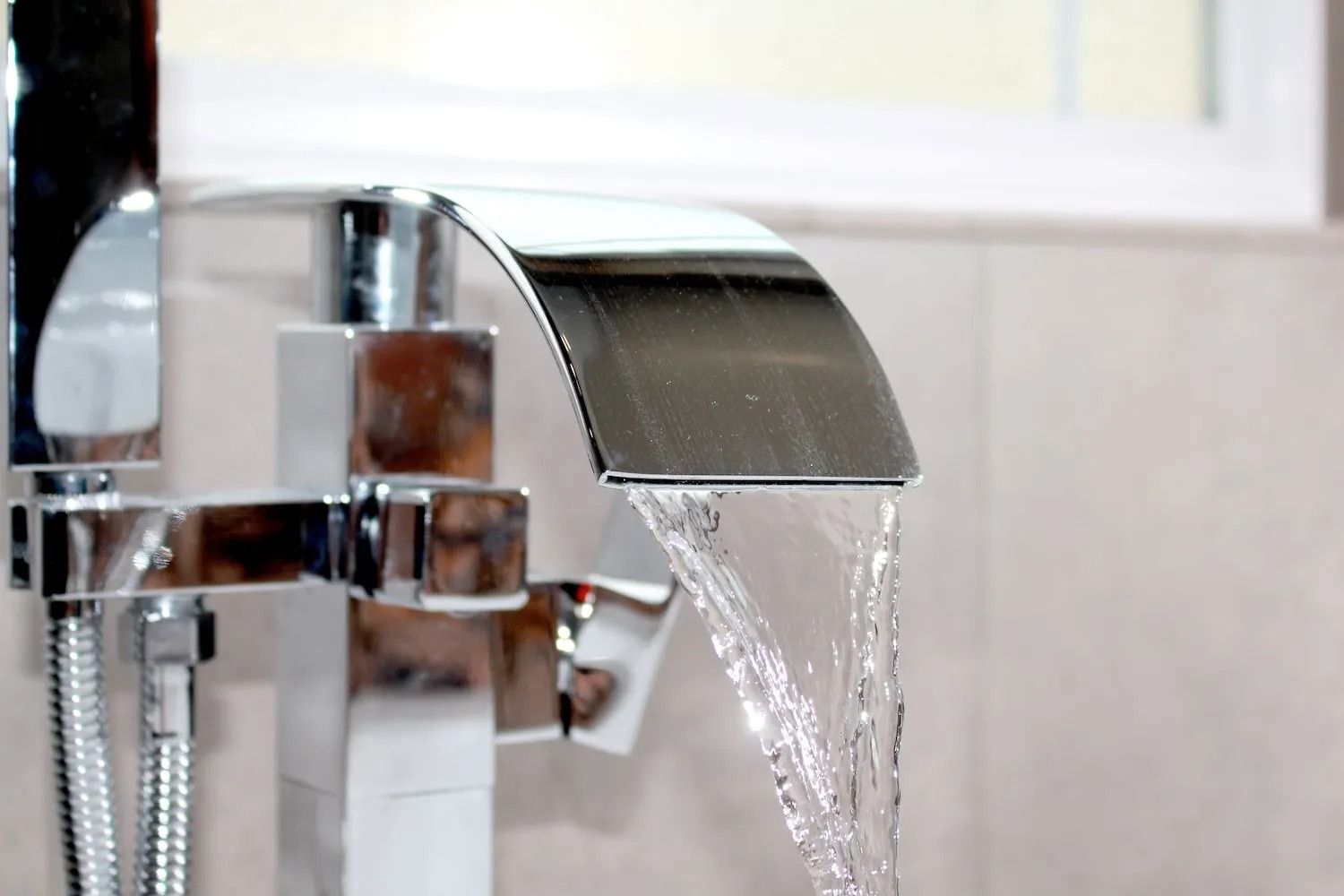A shiny chrome waterfall-style faucet pouring water into a basin against a tiled background.
