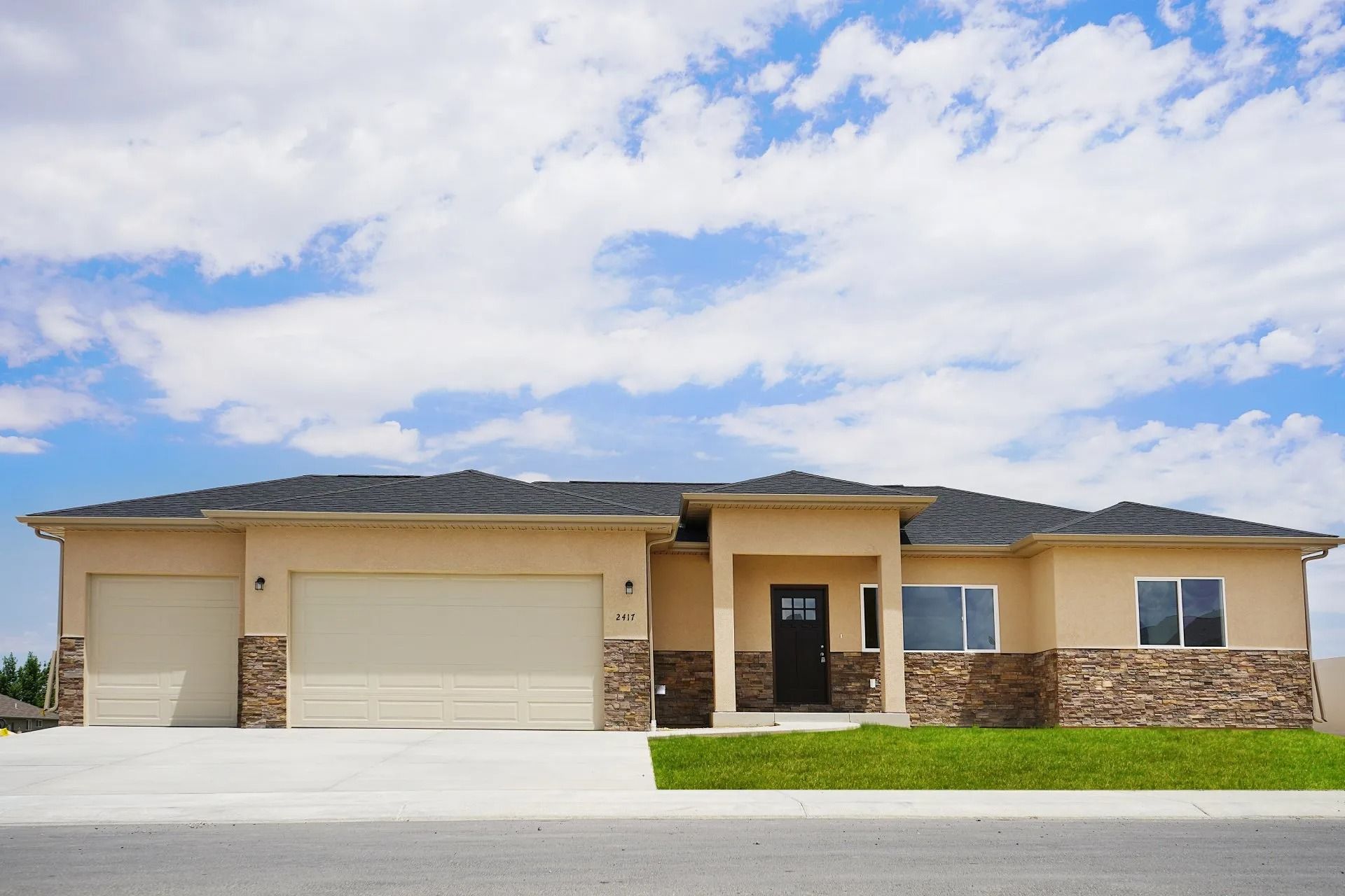 A single-story, beige stucco home with a three-car garage, stone trim, and a dark shingled roof under a blue sky.