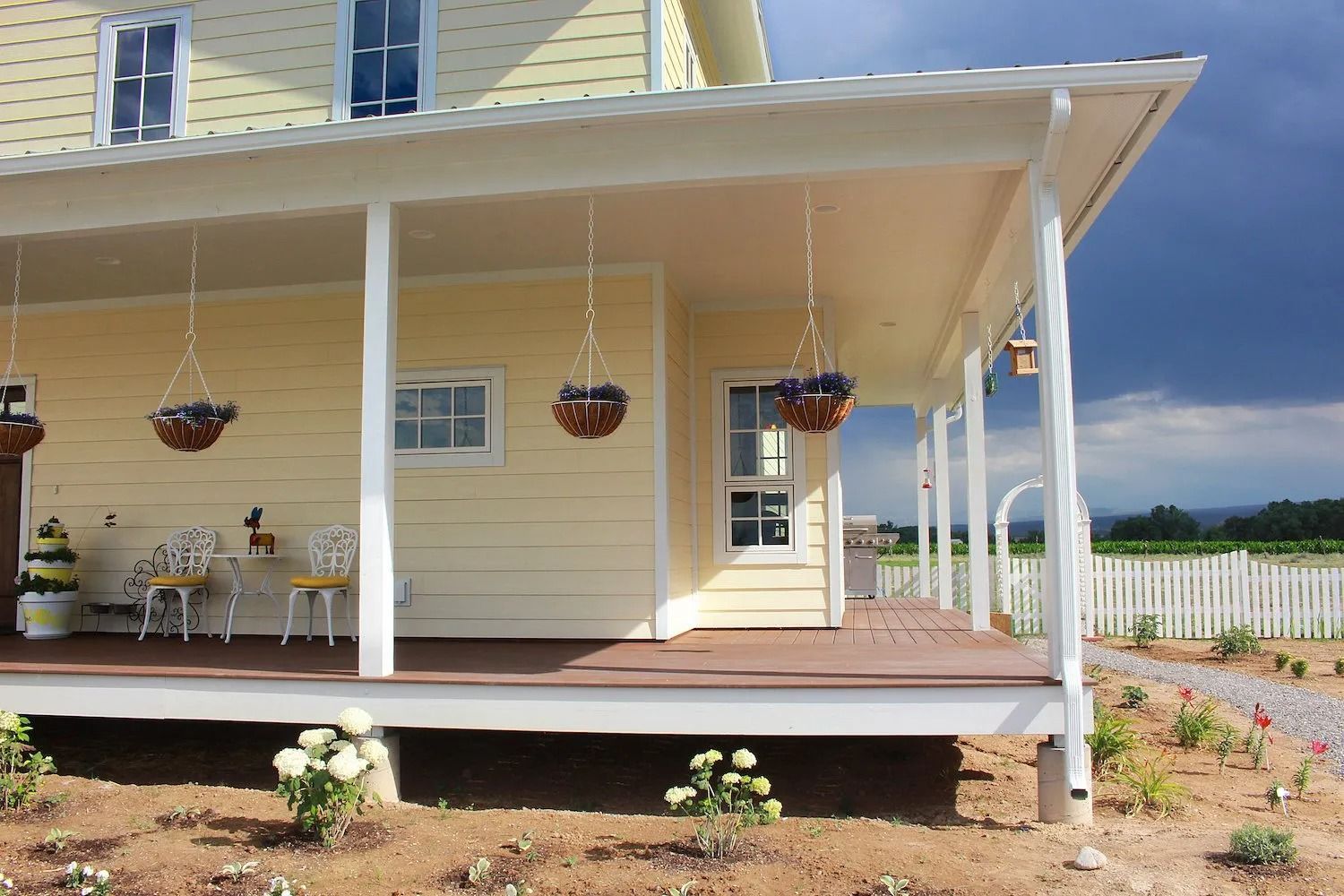 A yellow farmhouse with a covered porch featuring hanging plants, patio chairs, and a white picket fence in the distance.