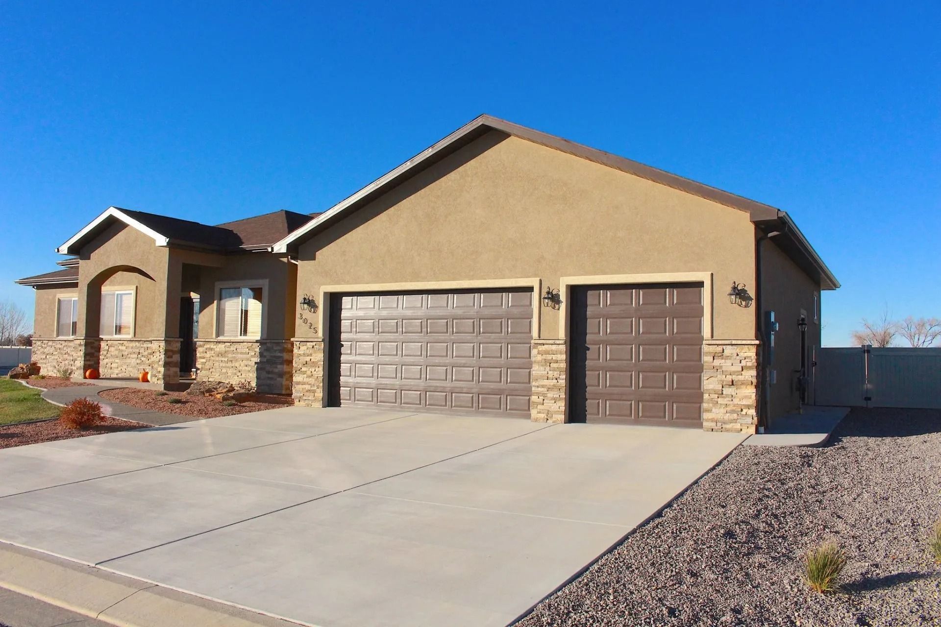A tan, single-story suburban house with stone accents, a concrete driveway, and a three-car garage under a blue sky.