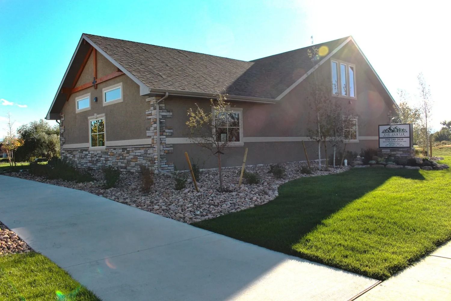 A single-story tan stucco and stone commercial building with a gray shingle roof under a bright sunny sky.