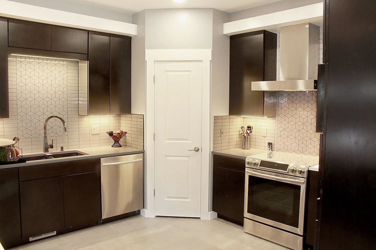 A modern kitchen with dark cabinetry, stainless steel appliances, a white door, and patterned tile backsplashes.