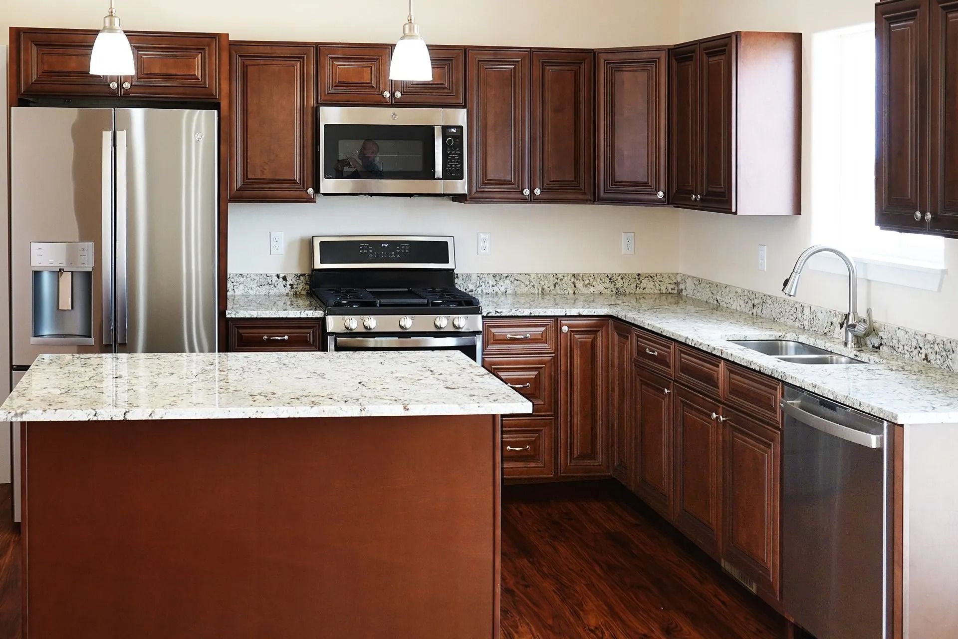 A modern kitchen featuring dark wood cabinets, white granite countertops, stainless steel appliances, and a central island.