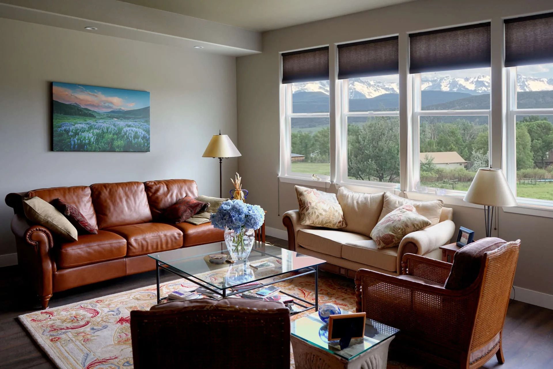 A living room with a brown leather sofa, tan loveseat, and wicker chair around a glass coffee table, facing a mountain view.