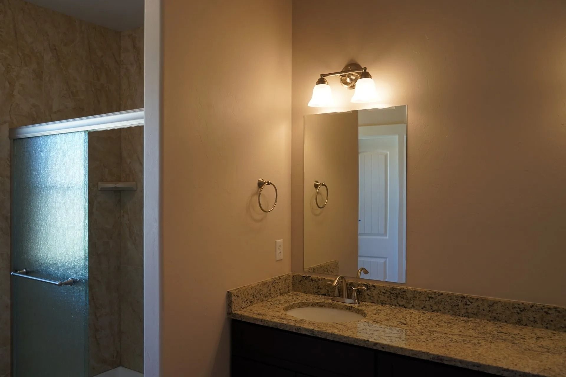 A bathroom with a dark vanity, speckled granite countertop, sink, wall mirror, two-light fixture, and a glass shower.