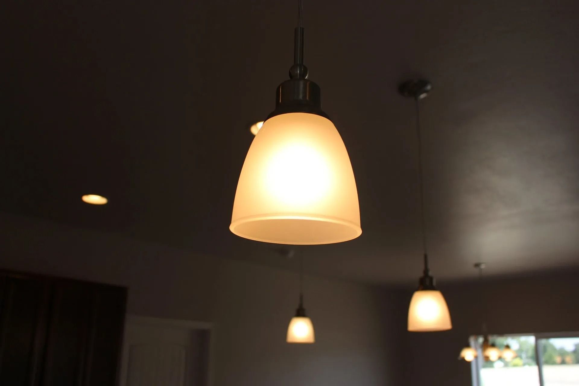 Four glowing, frosted glass pendant lights hang from a dark ceiling in a room.