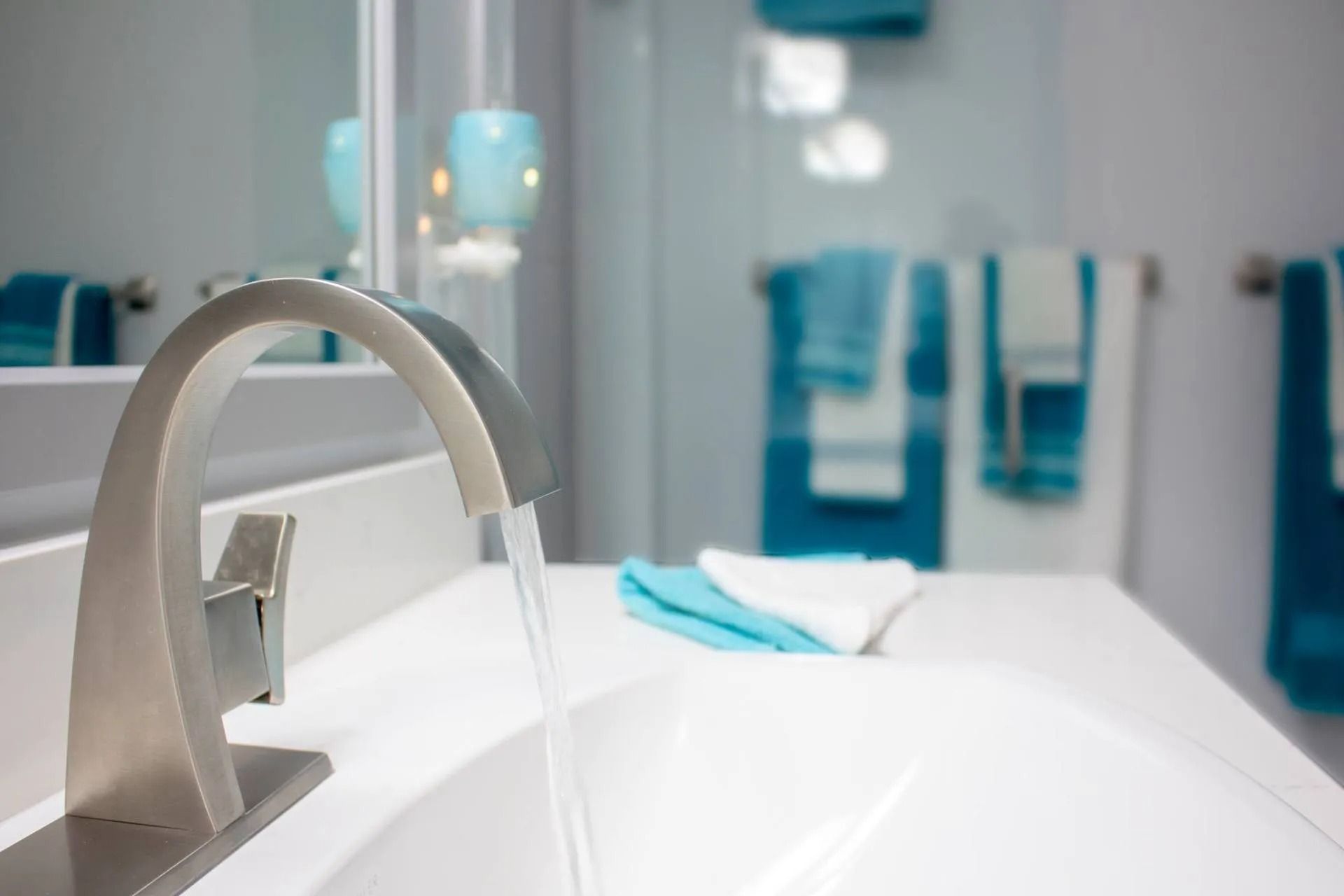 A modern silver bathroom faucet pours water into a white sink, with teal and white towels hanging in the blurred background.