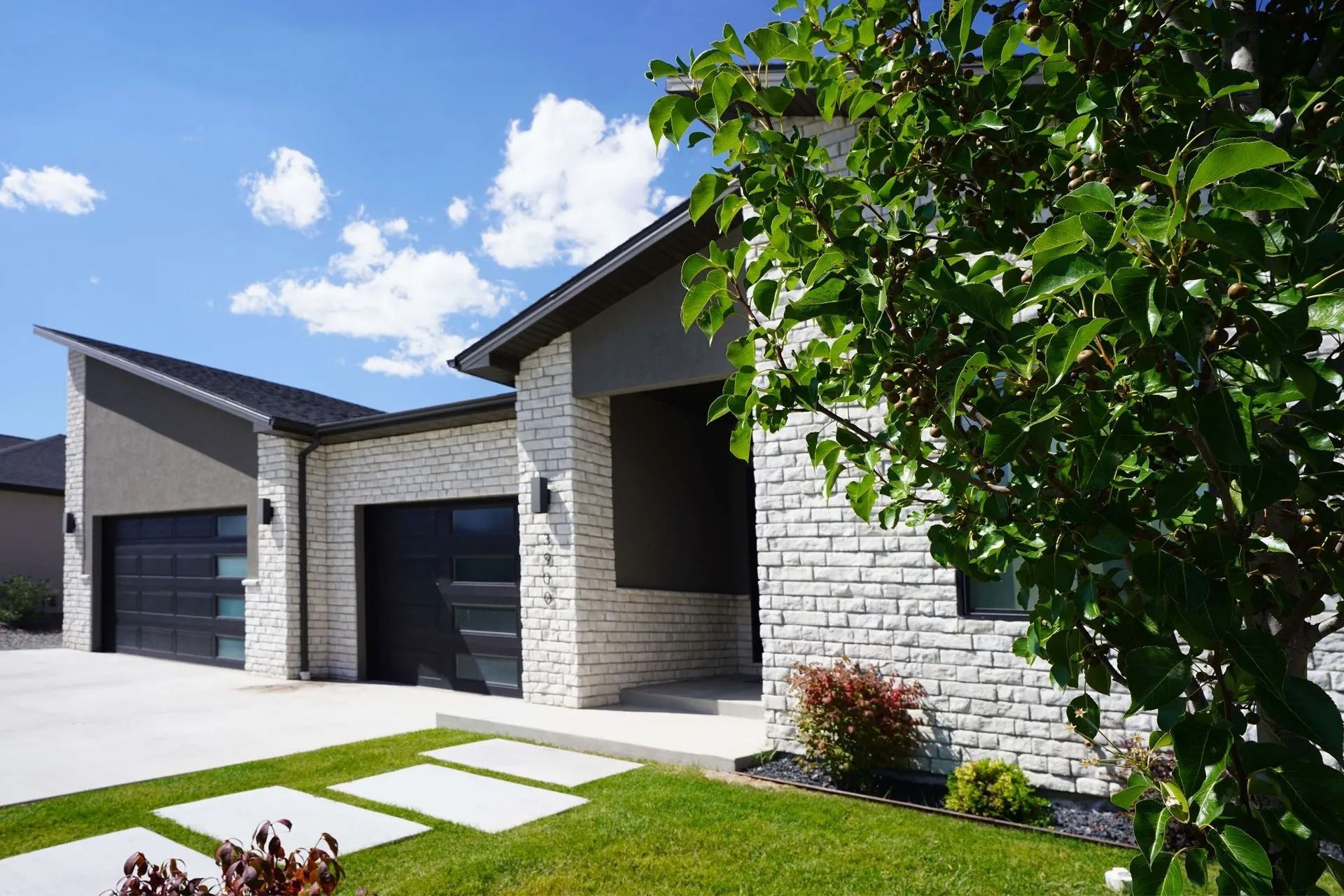 A modern house exterior with white stone siding, dark garage doors, and a green front lawn under a blue sky.