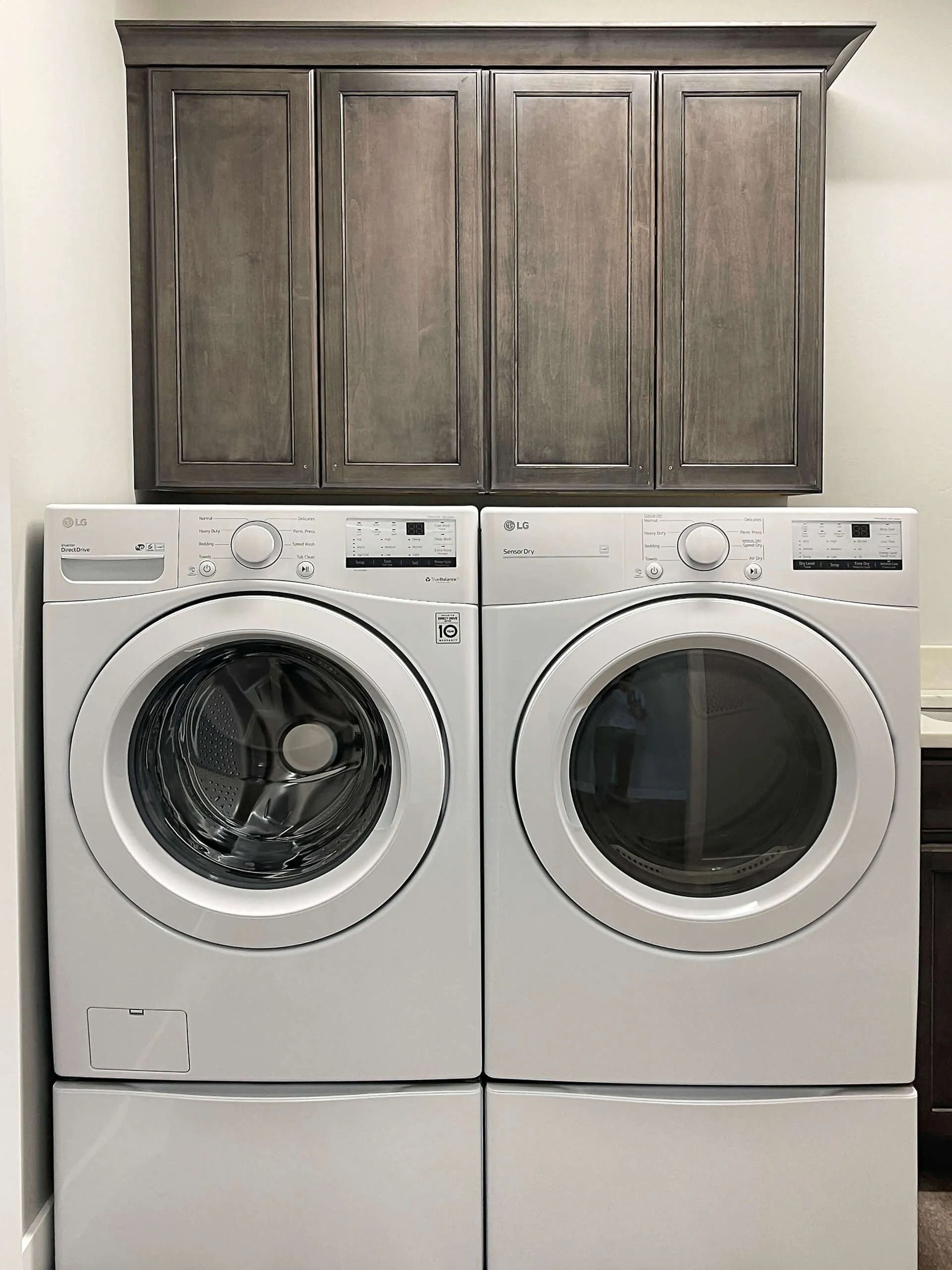 A side-by-side white washer and dryer set with matching storage pedestals beneath and a gray wall cabinet above.