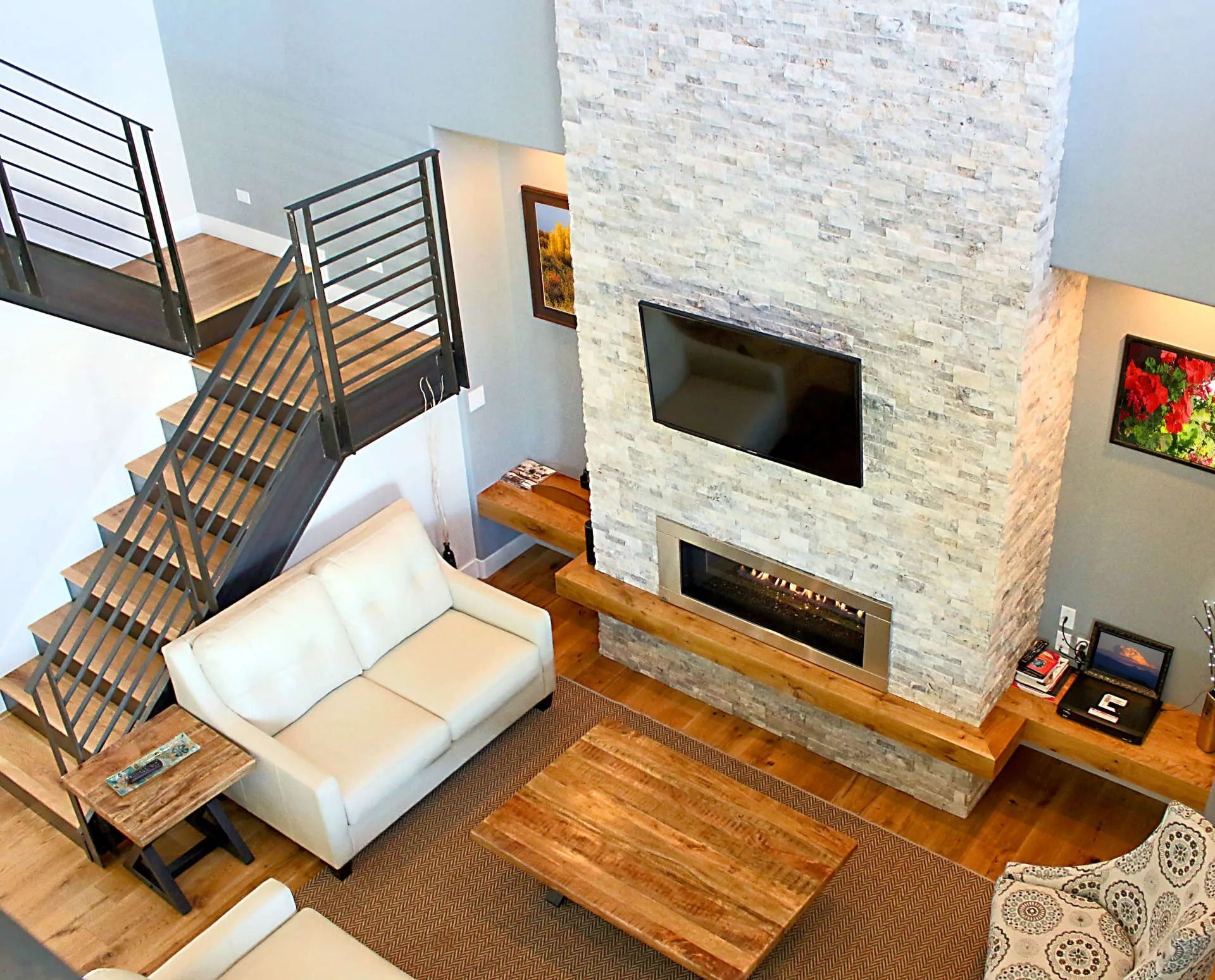 An elevated view of a living room with a white sofa, a wooden coffee table, a stone fireplace with a TV, and stairs.