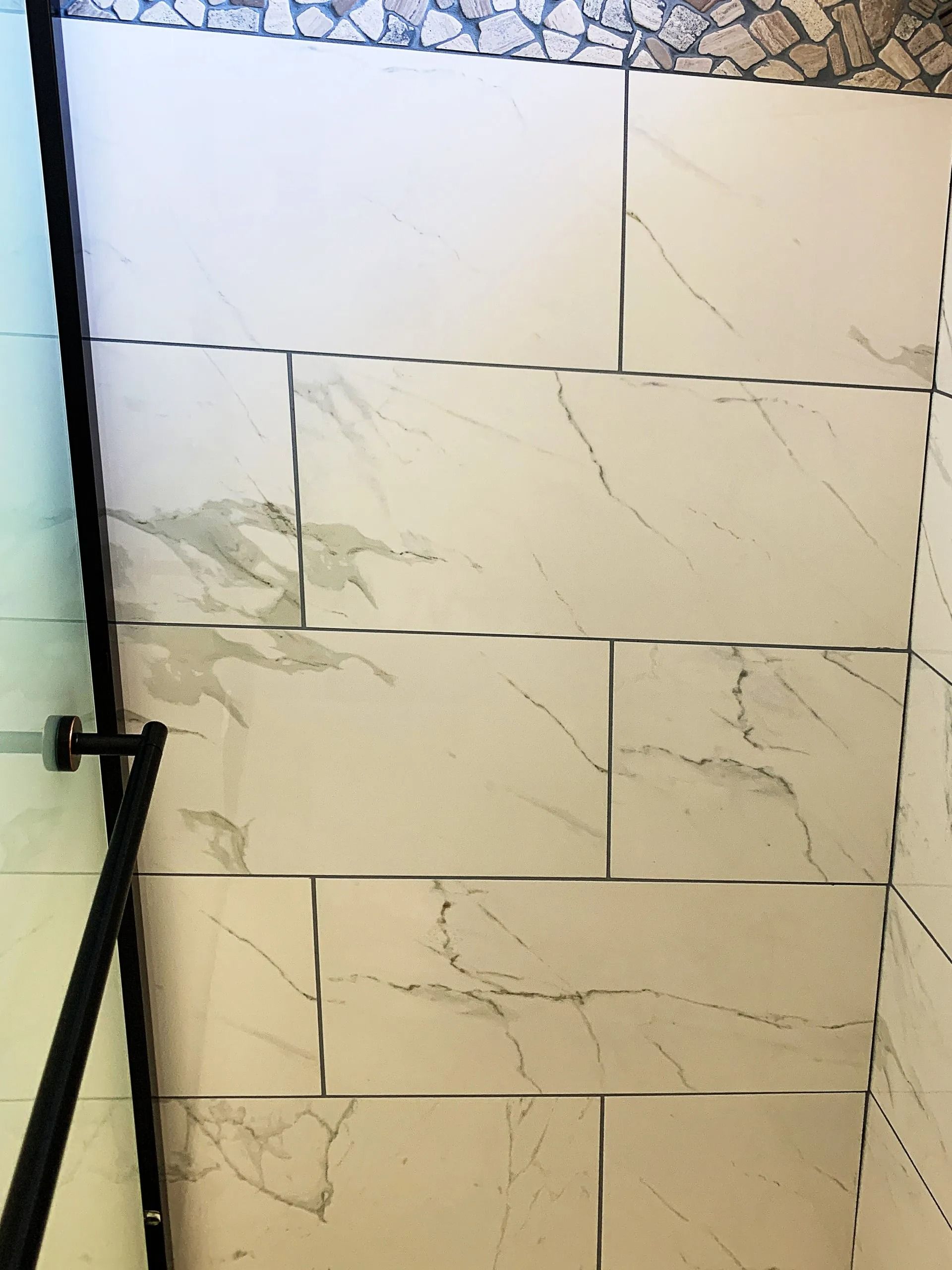 Close-up of a shower wall with large, rectangular, white marble-patterned tiles in a running bond pattern.