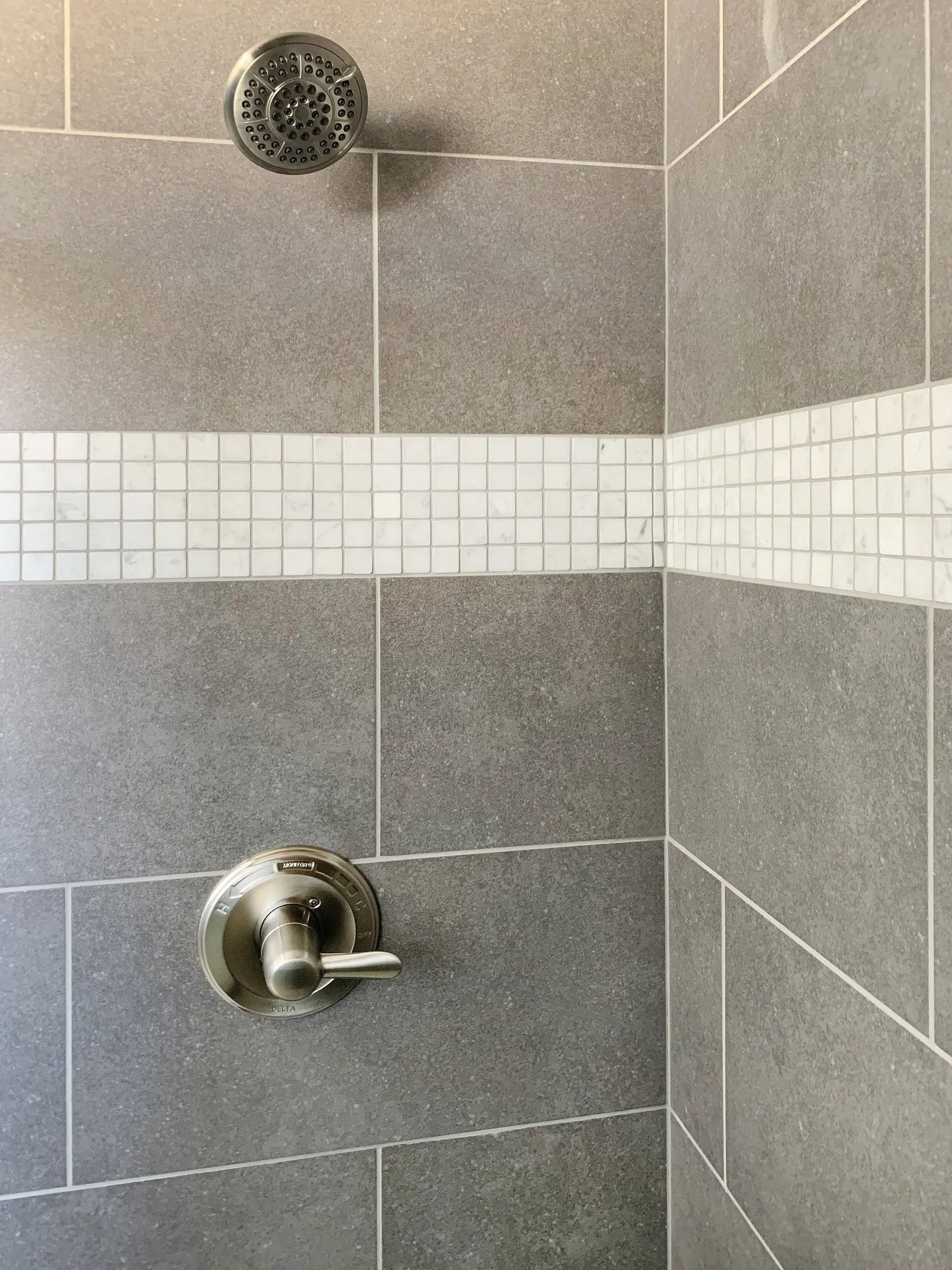 A shower wall with gray large-format tiles, a horizontal white mosaic accent strip, and a chrome shower head and handle.