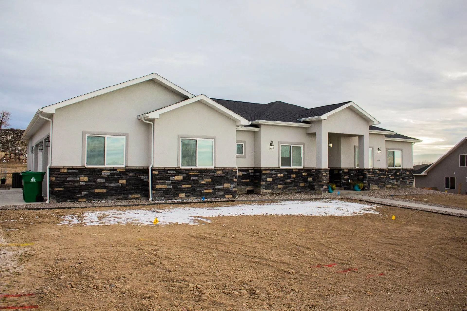 A single-story house with white stucco walls and dark stone siding sits on a plot of land with patches of snow.