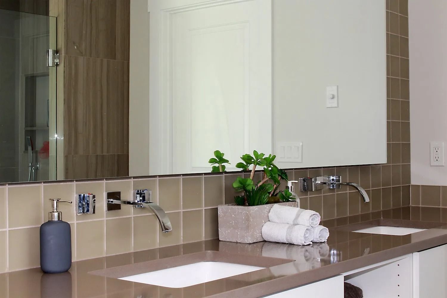 Modern bathroom vanity with double sinks, taupe square tiles, a grey soap dispenser, and a potted plant between faucets.