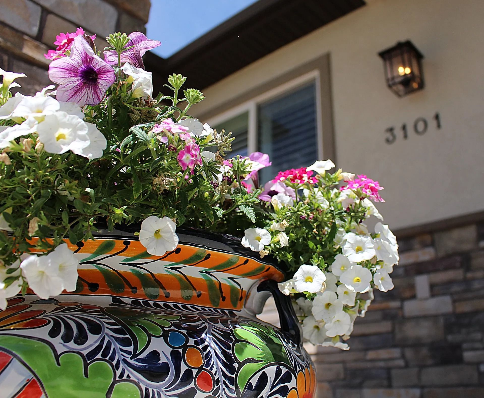 A colorful, hand-painted ceramic planter filled with white and pink petunias in front of a stone-faced house.