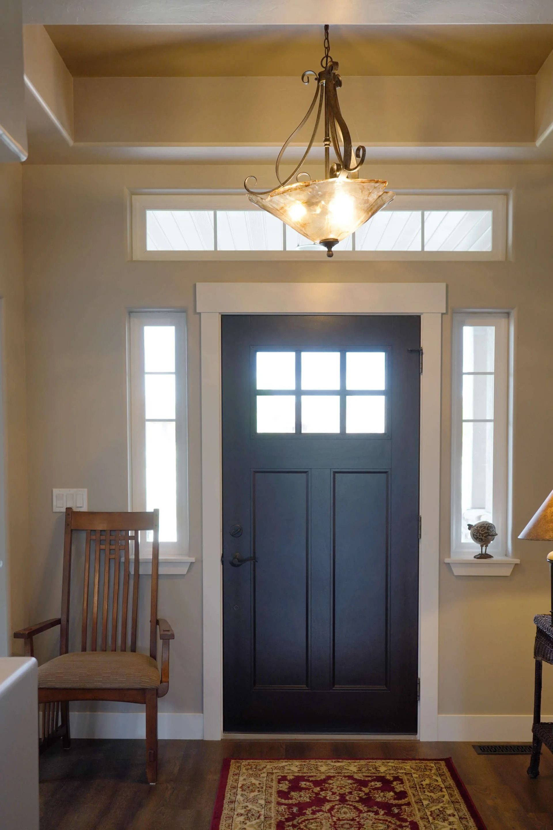A dark grey door with a glass pane, flanked by vertical windows and a transom, with a wooden chair in an entryway.