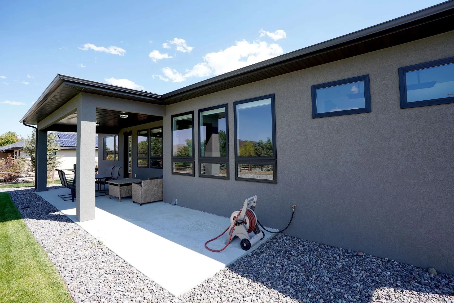 A patio with furniture next to a gray stucco house with large windows and a garden hose reel on a sunny day.