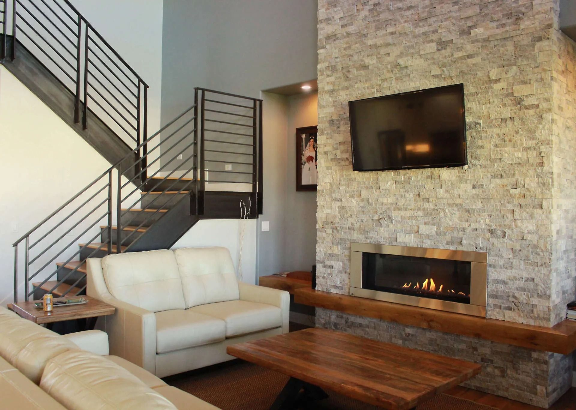 A modern living room with a stone fireplace, wall-mounted television, wooden coffee table, sofa, and a staircase.