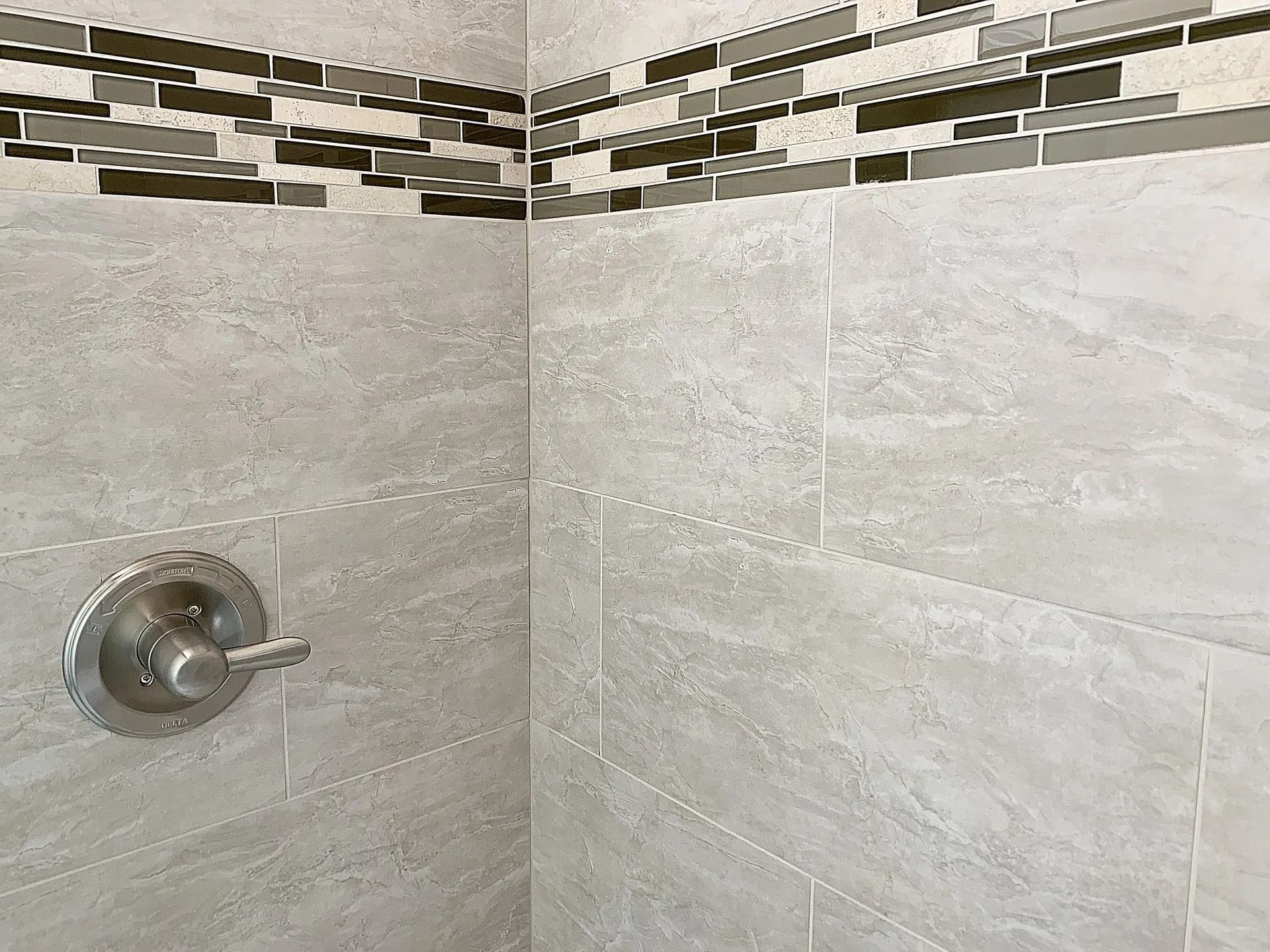 A shower wall with gray stone-look tiles and a horizontal decorative accent border, featuring a metal shower control valve.