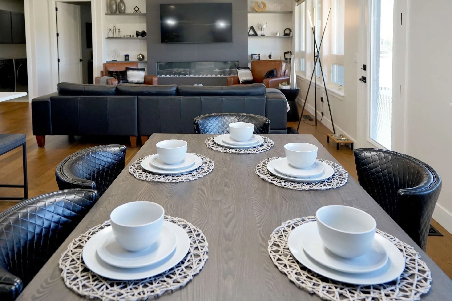 A modern dining table with four place settings, featuring white bowls and plates on textured mats, with a living room.