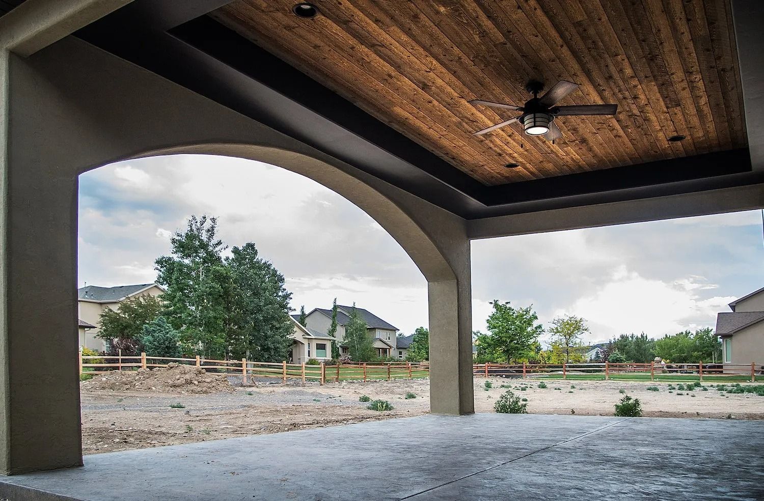 A covered patio with a wooden ceiling, ceiling fan, and arched opening looking out over a landscaped suburban yard.