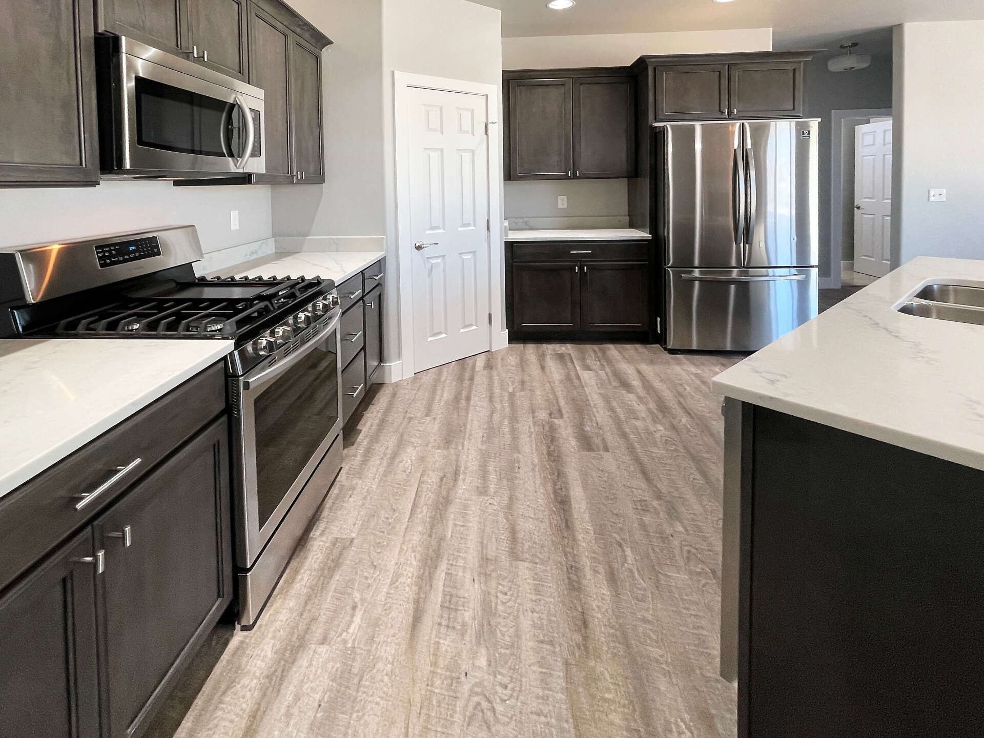 Modern kitchen with dark wood cabinets, stainless steel appliances, white countertops, and light wood-look vinyl flooring.