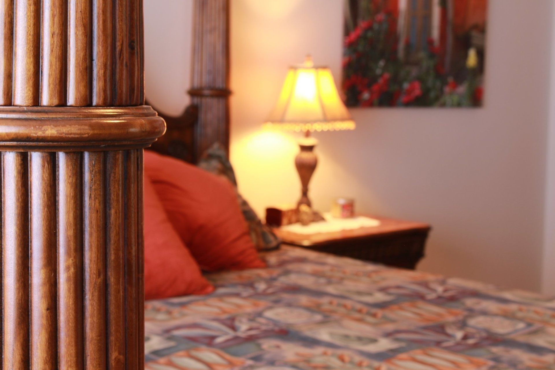 A close-up of a fluted wooden bedpost in the foreground, with a blurred bed, red pillows, and a lit table lamp behind it.