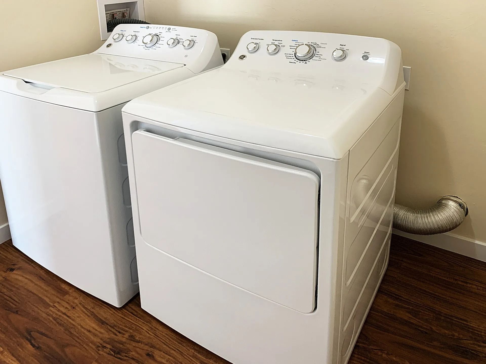 A white clothes washer and a matching dryer with a silver vent pipe on a wooden floor against a beige wall.