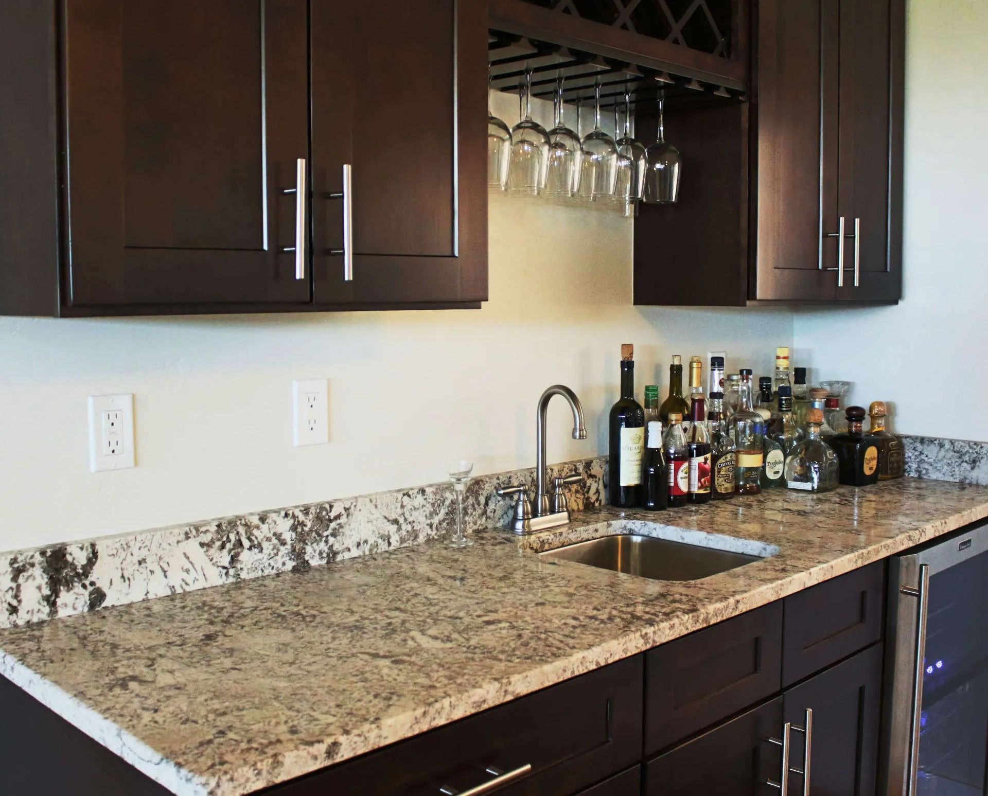 A home wet bar with dark wooden cabinets, granite countertops, a stainless steel sink, wine glass rack, and alcohol bottles.