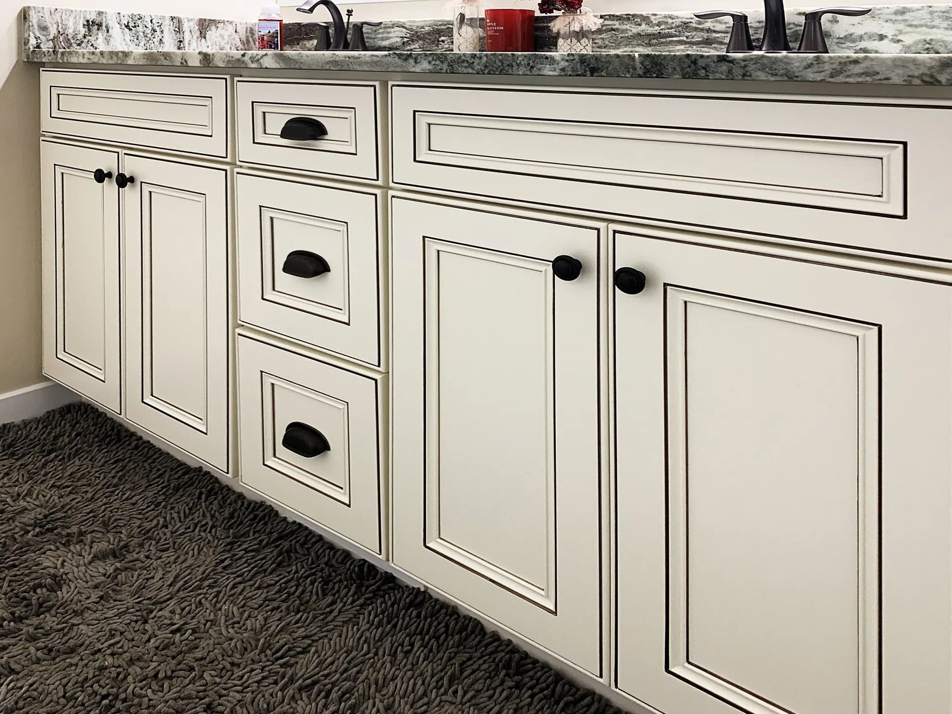 Off-white bathroom vanity cabinets with black trim, dark hardware, and a marble countertop sitting on dark carpet.