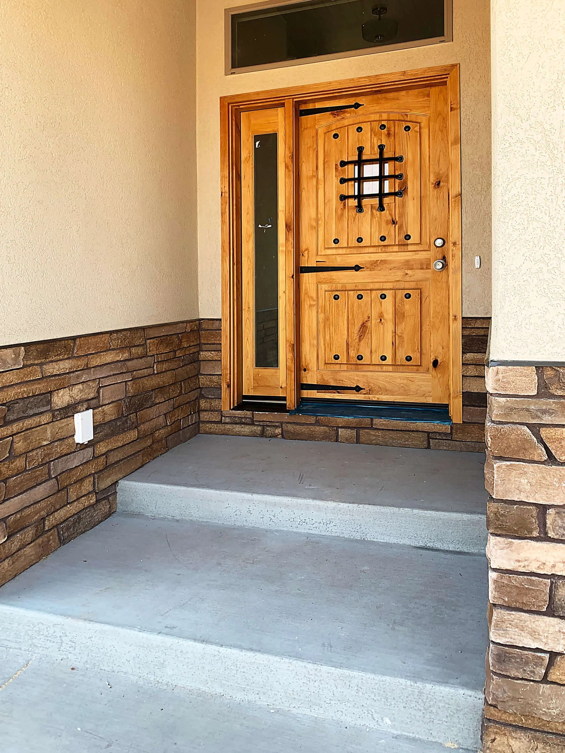 Rustic wooden front door with a metal-grate window and side light, set into a stone-veneer entryway with concrete steps.