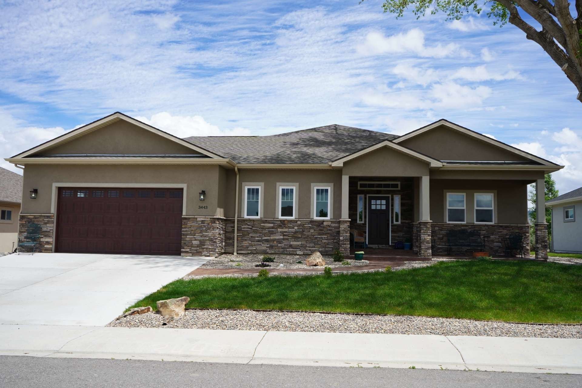 A single-story suburban house with brown stucco walls, stone accents, a dark garage door, and a grass front lawn.