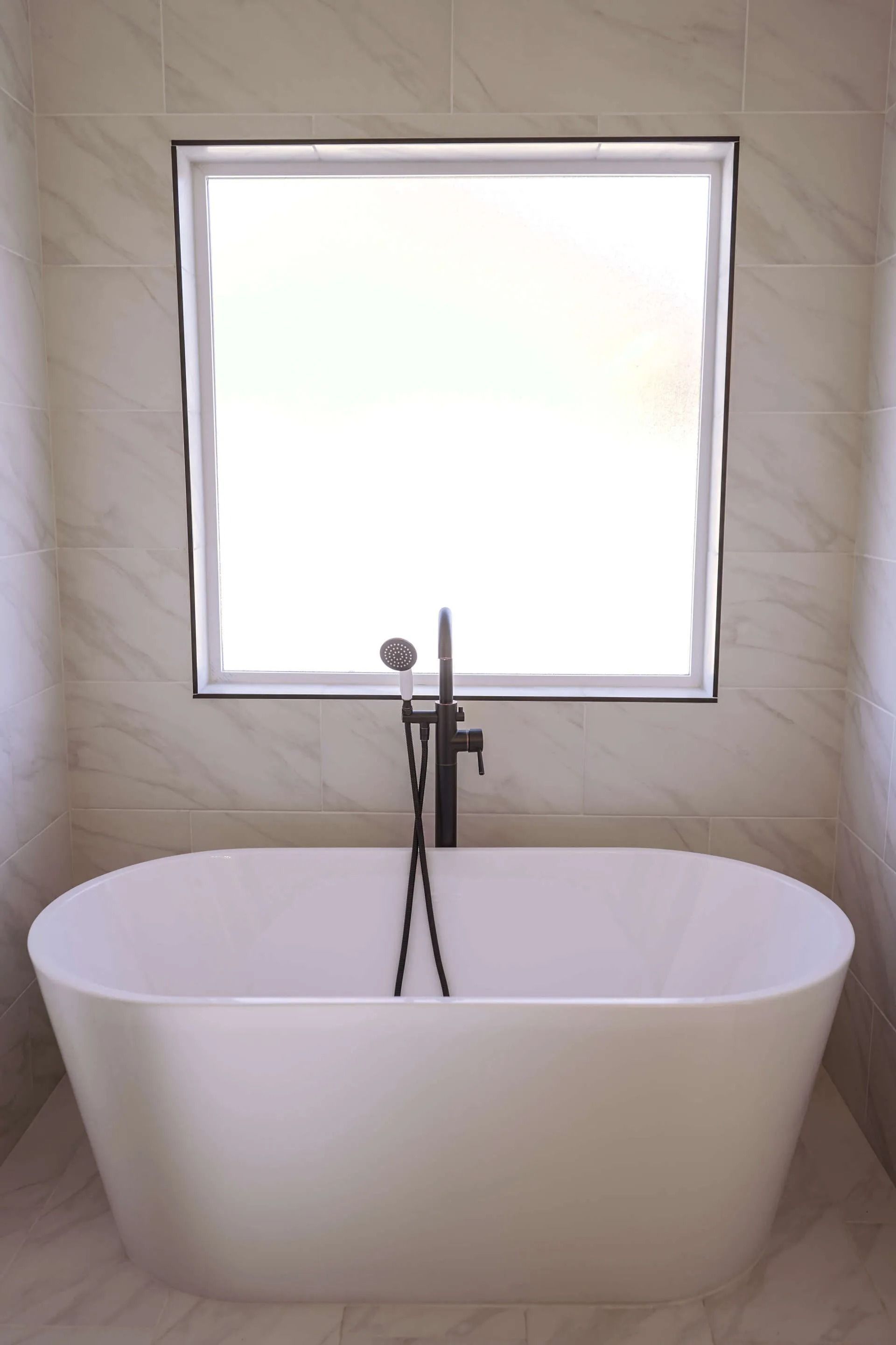 A modern white freestanding bathtub placed in front of a square window against a light-colored tiled wall.