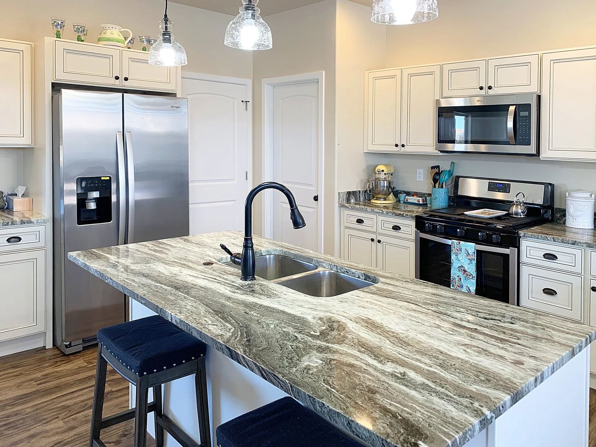A modern kitchen with white cabinets, stainless steel appliances, a large granite-topped island, and two navy stools.