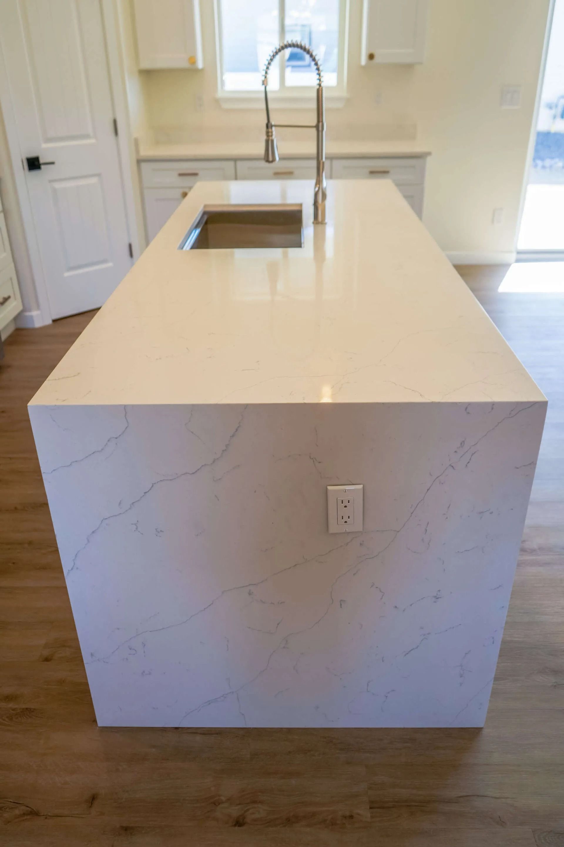 A white quartz kitchen island with gray veining features a center sink, a tall faucet, and a wall outlet on the end.