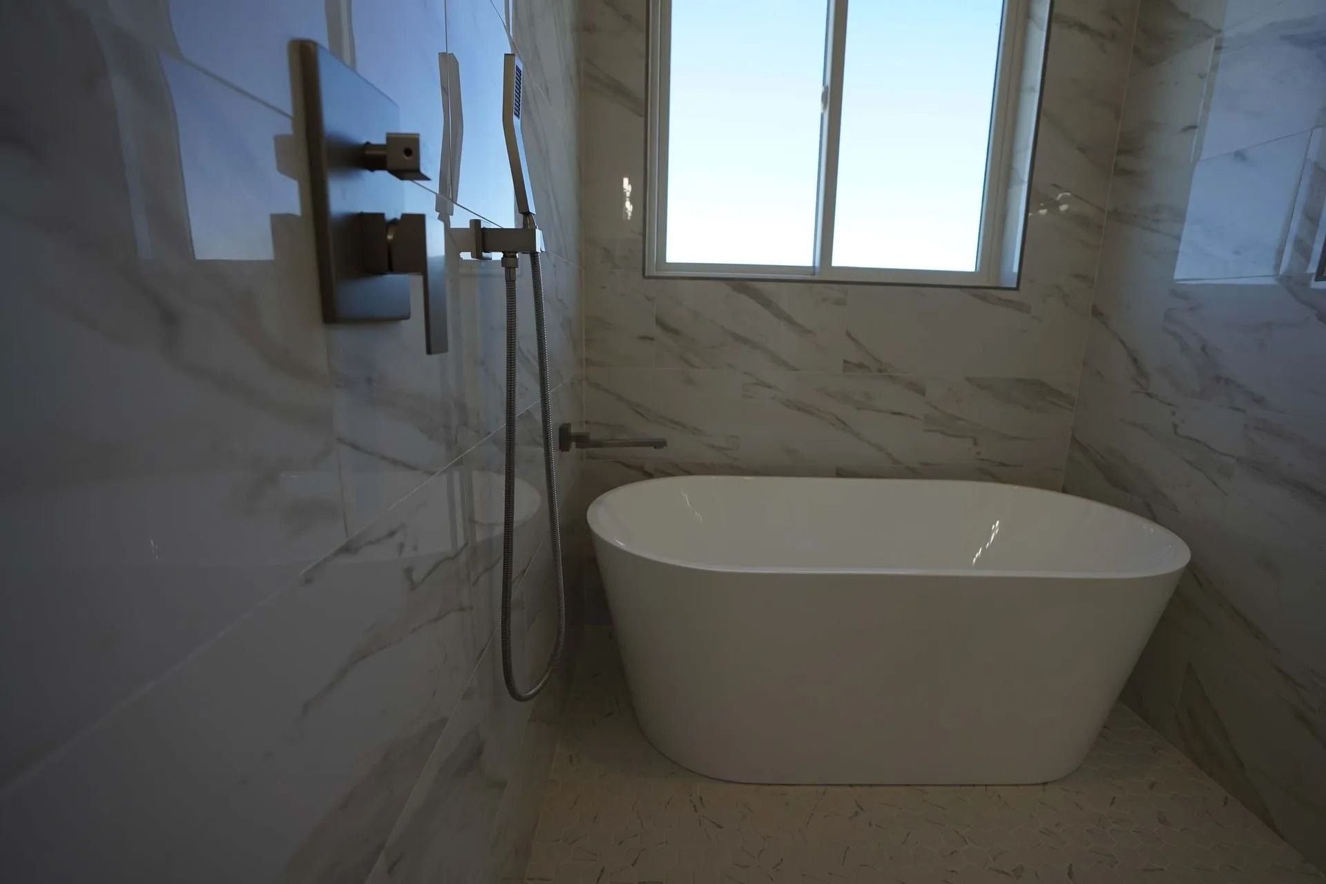 A white freestanding bathtub sits in a room with light-colored marble tile walls and a window above the tub.