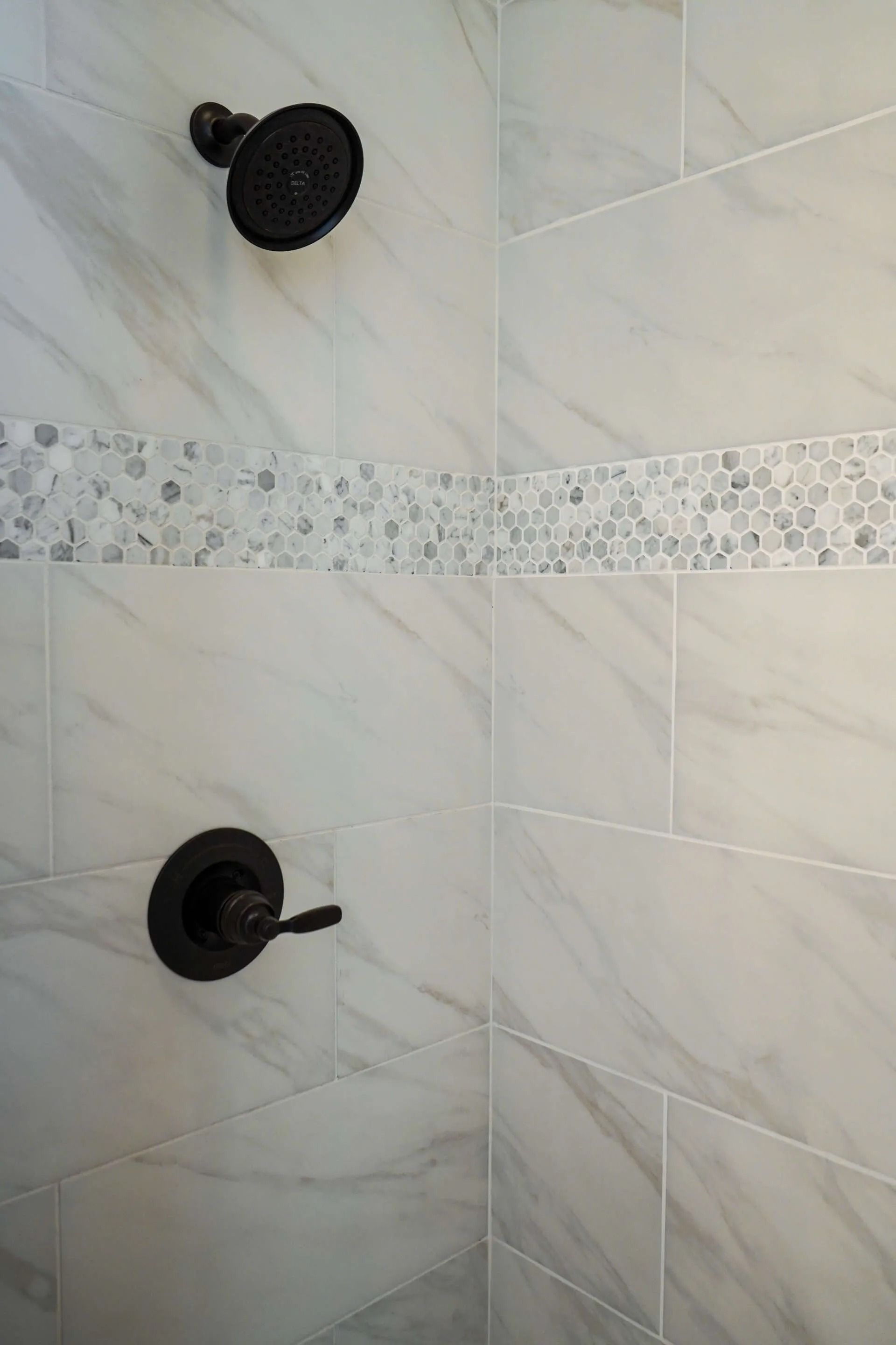 A shower with white marble-style wall tiles, a horizontal pebble inlay border, and black fixtures.
